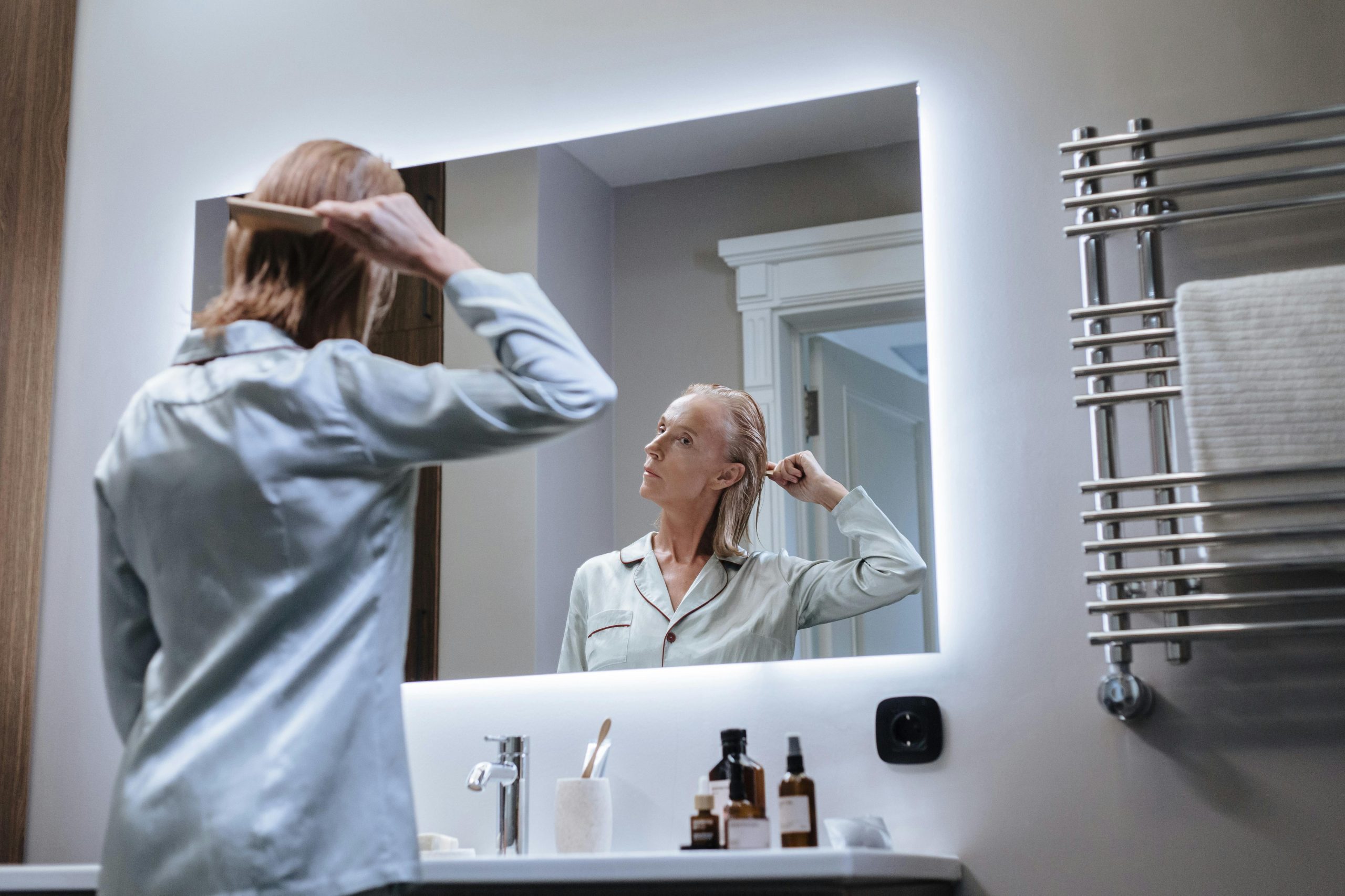 Woman in her 40s looking in her bathroom mirror, taking stock — wellness products on the counter
